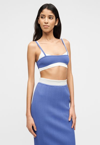 Jeune femme portant un haut court en maille bleu et blanc et une jupe côtelée taille haute assortie, debout devant un fond uni.