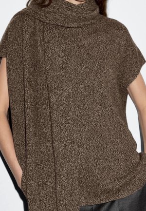 Femme portant un haut en tricot marron sans manches avec une longue écharpe assortie drapée autour du cou et un pantalon foncé, debout les mains dans les poches.