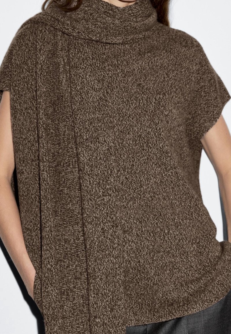 Femme portant un haut en tricot marron sans manches avec une longue écharpe assortie drapée autour du cou et un pantalon foncé, debout les mains dans les poches.