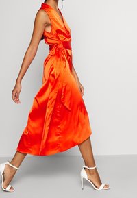 Pomarańczowa sukienka kopertowa z satyny z głębokim dekoltem w kształcie litery V i paskiem do wiązania. Charakteryzuje się zwiewną długością do połowy łydki. Noszona z białymi sandałami na wysokim obcasie z paskami.