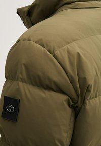 Oliwkowa kurtka puchowa z pikowanym wzorem, wysokim kołnierzem i czarną łatą z logo na ramieniu. Gładki materiał, ciepła izolacja.