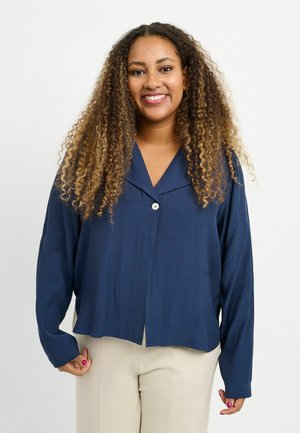 Lachende vrouw met lang krullend haar, gekleed in een marineblauw jasje met knopen en beige broek, staand tegen een effen witte achtergrond.