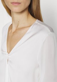 Bílá blůza s výstřihem do V a nařasením vpředu, vyrobená z lehké, hladké tkaniny a s dlouhými rukávy.