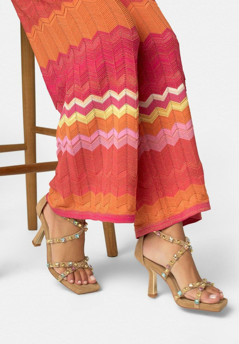 Femme portant un pantalon à motif zigzag multicolore assise sur un tabouret en bois, sandales à talons beiges avec des lanières ornées de pierres précieuses colorées aux pieds.