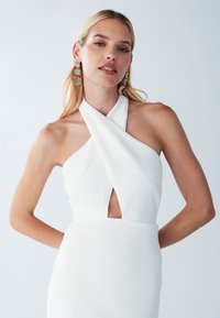 Weißes Neckholderkleid mit einem Front-Ausschnitt, ausgestattet mit einem strukturierten Stoff. Das Modell trägt große transparente Ohrringe und hat glattes Haar.