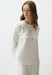 Witte lange mouwen top met een ronde halslijn, versierd met accenten die lijken op rhinestones, gerangschikt in bloemmotieven. Zachte stof textuur.