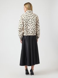 Γυναίκα με ξανθά μαλλιά που φοράει μπεζ σακάκι με λευκά πουά, μαύρη μίντι φούστα και μαύρα ψηλοτάκουνα παπούτσια, στέκεται γυρισμένη προς τα πίσω σε λευκό φόντο.