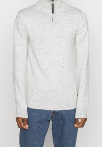 Maglione grigio chiaro con colletto alto, chiusura a mezzo zip, polsini a coste e un motivo testurizzato di macchie più scure.
