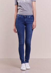 Smala blå denimjeans med midjehög passform, fem fickor och en subtil blekt finish, tillsammans med en grå t-shirt med tryck och vita sneakers.