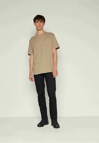 T-shirt beige a maniche corte con dettaglio tasca, abbinato a pantaloni slim-fit neri e scarpe nere su uno sfondo neutro.