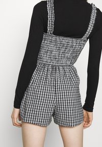 Randig jumpsuit i svart och vitt med en åtsittande liv, volangiga axelband och elastisk midja, kombinerad med en långärmad svart topp.