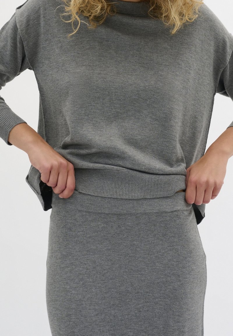 Femme en pull gris à manches longues et jupe assortie, ajustant l'ourlet du pull avec les deux mains sur un fond blanc uni.