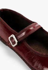 Zapato Mary Jane de charol burdeos con un acabado texturizado, correa de tela y una hebilla decorativa plateada. Forma clásica redondeada.