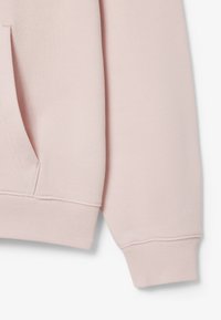 Tela de sudadera rosa claro con un acabado texturizado, que presenta un bolsillo lateral y puños acanalados. Detalles de costura visibles a lo largo de las costuras.