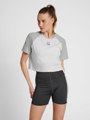 Beskåret hvid t-shirt med grå raglanærmer, prydet med et lilla logo. Sammen med tætsiddende sorte atletikshorts med sidelommer.