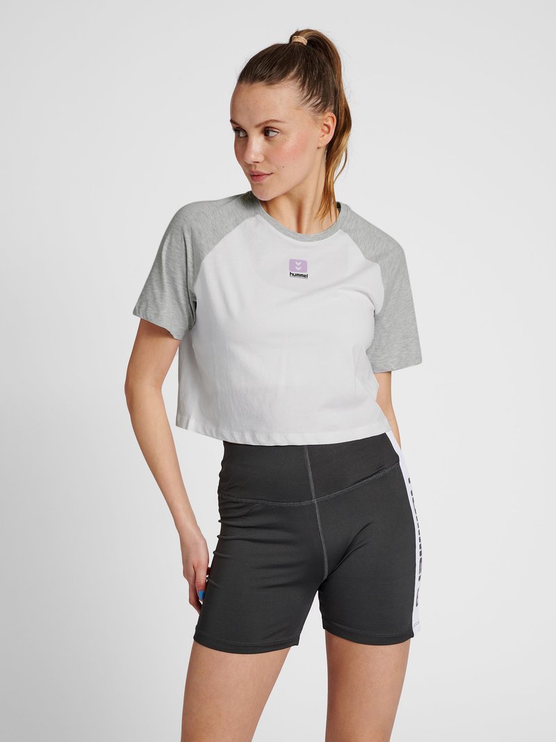 Abgeschnittenes weißes T-Shirt mit grauen Raglanärmeln, versehen mit einem lila Logo. Kombiniert mit enganliegenden schwarzen Sportshorts mit seitlichen Akzenten.