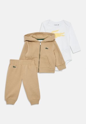 Beežid imikute dressipüksid, lukuga kapuutsiga jakk ja valge pika varrukaga bodi kollase krokodillilogoga rinnal.