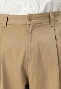 Beige trousers with a smooth texture, button closure, and pleated front design. Features side pockets and sturdy stitching.