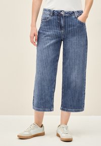 Culottes de mezclilla en azul con rayas verticales blancas, con una cinturilla distintiva, cinco bolsillos y dobladillo acabado. Combinados con zapatillas ligeras.