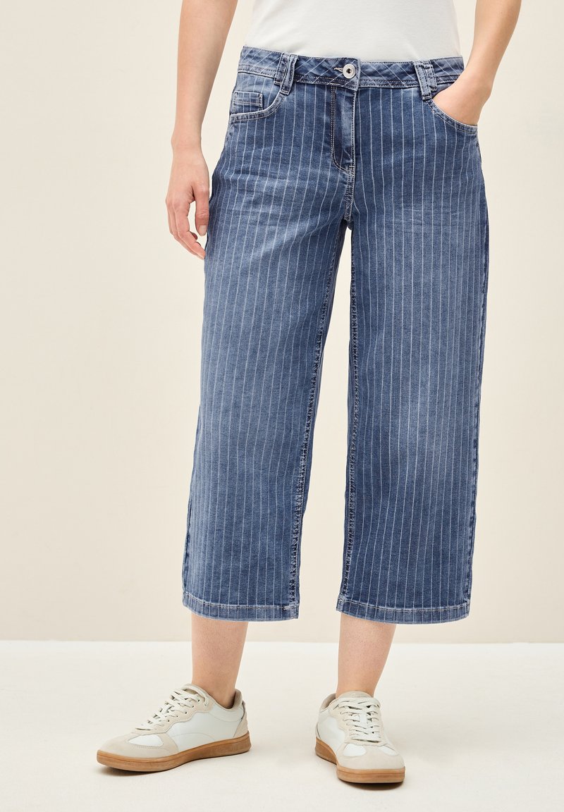 Culottes de mezclilla en azul con rayas verticales blancas, con una cinturilla distintiva, cinco bolsillos y dobladillo acabado. Combinados con zapatillas ligeras.