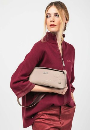 Frau in einem weinroten Zip-up-Oberteil und passenden Hosen, die eine beige Handtasche unter dem Arm hält und zur Seite blickt, vor einem einfarbigen Hintergrund.