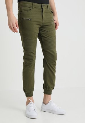 Homme portant un pantalon de jogging vert olive avec des poches zippées et un détail côtelé aux genoux, associé à des baskets blanches et un haut noir.