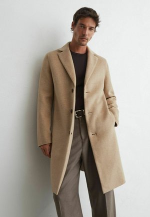 Man wearing a beige long coat, dark shirt, and brown trousers, standing against a white textured wall with one hand in his pocket.