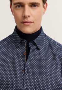 Camicia button-up a motivi blu navy con un design geometrico, bottoni marroni e un colletto scuro a contrasto. Tessuto di peso medio con una consistenza liscia.
