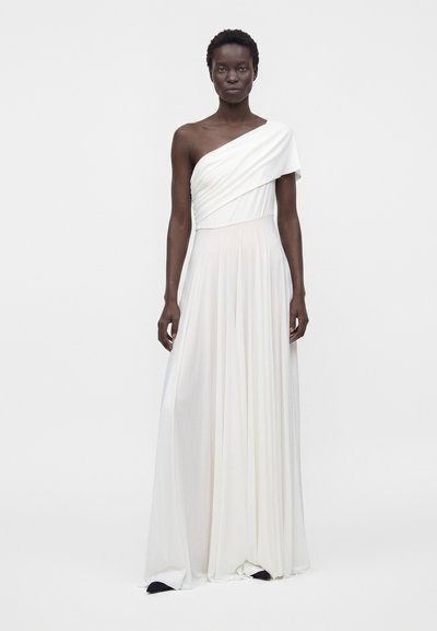 Mujer con un vestido blanco de un solo hombro, largo hasta el suelo y fluido, de pie frente a un fondo liso de color gris claro.