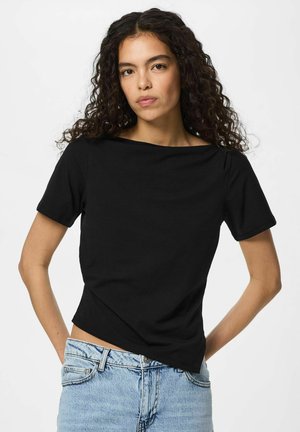 Nuori nainen, jolla on kiharat tummat hiukset, pukeutunut mustaan lyhythihaiseen toppiin ja vaaleansinisiin farkkuihin, seisoo käsivarret lanteilla valkoista taustaa vasten.