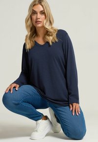 Navy-blaues Langarmoberteil mit V-Ausschnitt und Verzierungen, getragen mit blauen Jeans und weißen Sneakern, vor einem hellen Hintergrund.