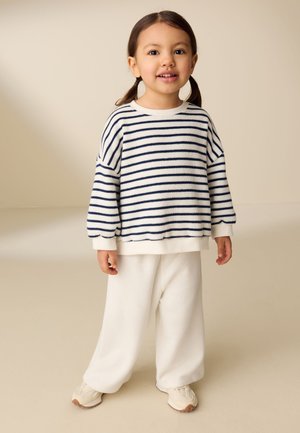 Lille barn med brunt hår i to fletninger iført en hvid og marineblå stribet sweater, hvide bukser og beige sneakers, står på en ensfarvet baggrund.