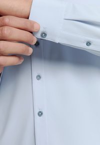 Lichtblauw overhemd met lange mouwen van een gestructureerde stof, voorzien van knopenmanchetten en bijpassende blauwe knopen. Close-up van de manchetdetails en knopen.
