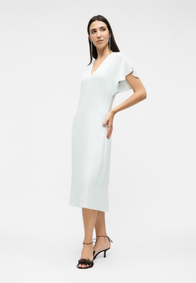 Γυναίκα με λευκό μίντι φόρεμα με φαρδιά μανίκια, φοράει μακριά σκουλαρίκια και μαύρα ψηλοτάκουνα σανδάλια, ποζάροντας με το χέρι στη μέση.