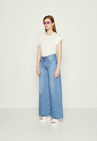 White top with ruffled sleeves, paired with wide-leg blue denim jeans and white platform shoes. Model wears purple sunglasses.