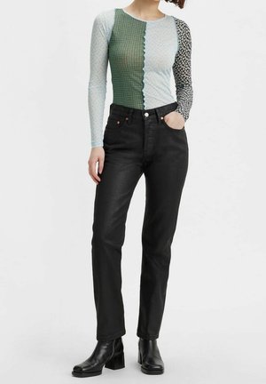 Femme portant un haut patchwork à manches longues avec des motifs variés, un jean noir taille haute et des bottines noires à talons.