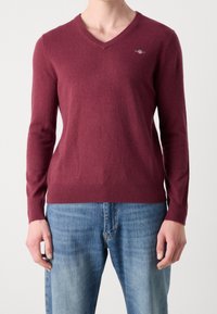 Pull col en V bordeaux en tissu tricoté doux, coupe ajustée avec manches longues, poignets et ourlet côtelés, présentant un petit logo sur la poitrine.