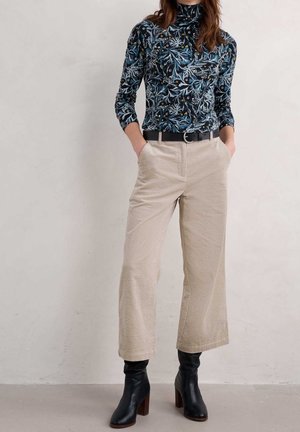 Woman wearing a dark floral turtleneck, beige wide-leg corduroy pants with black belt, and black heeled ankle boots, standing with hands in pockets.