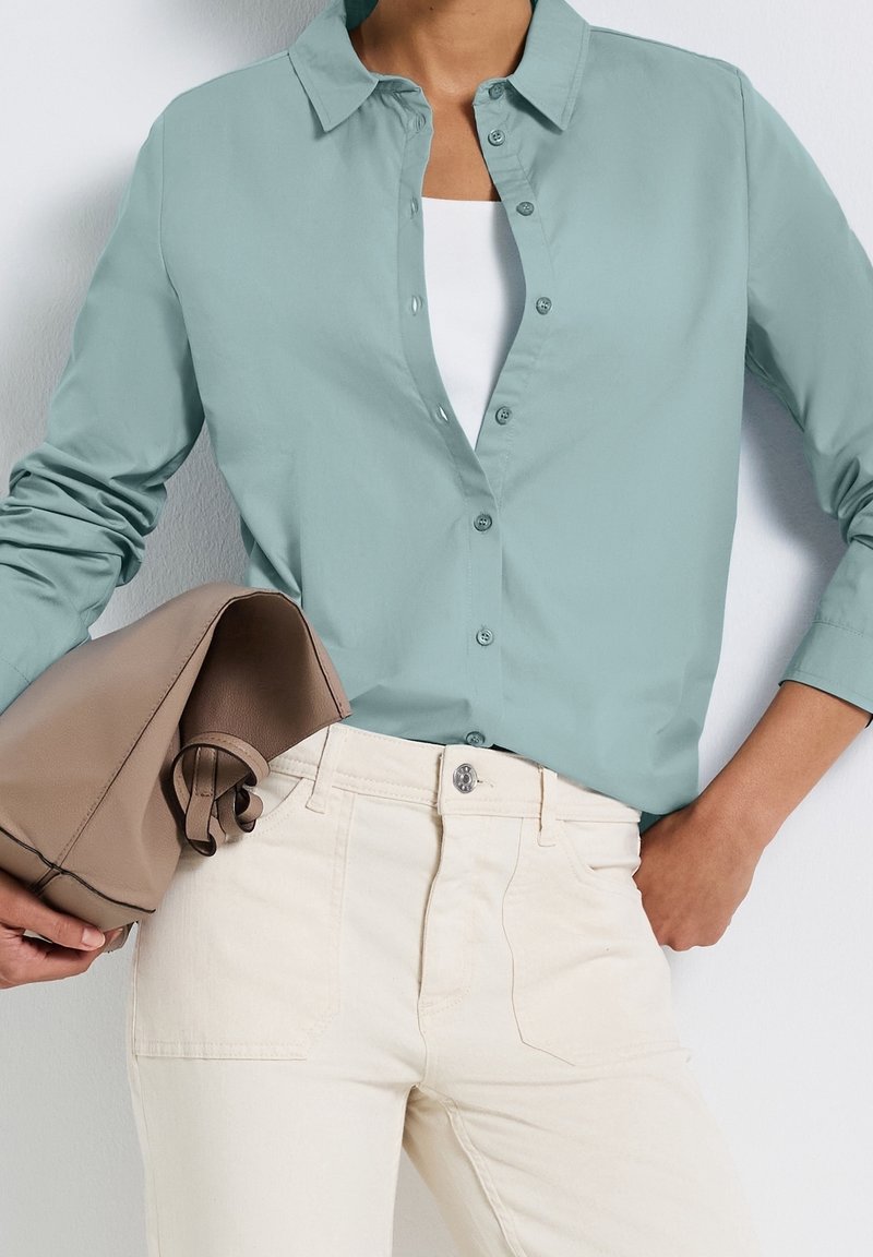 Personne portant une chemise bleu clair boutonnée sur un haut blanc, un pantalon crème, tenant un sac à main beige plié, une main dans la poche.