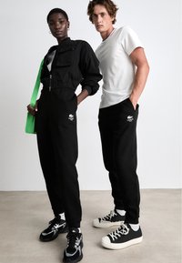 Zwarte cropped jack met rits, zwarte sweatpants met logo, witte t-shirt, groene schoudertas en contrasterende sneakers in zwart en wit.