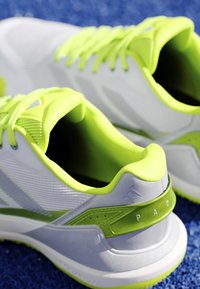 White and neon green sports shoes placed on a blue textured surface, showing the rear and side details.