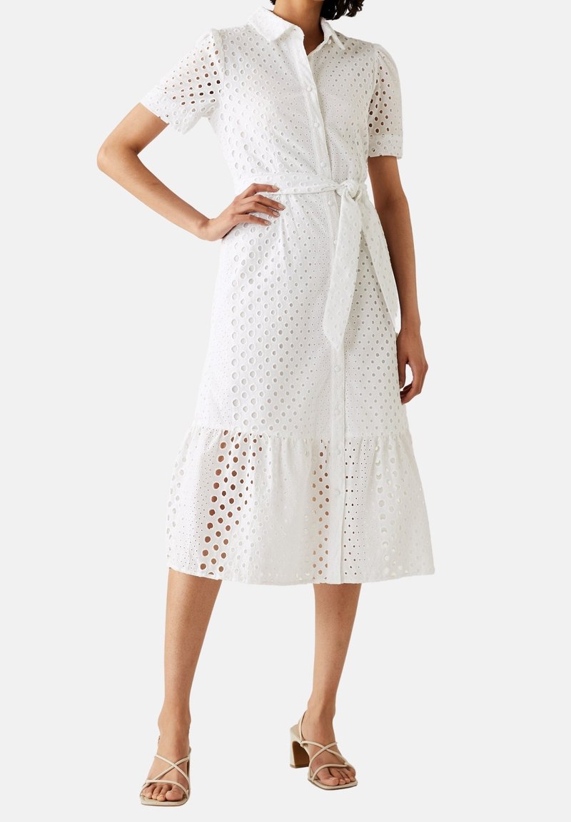 Biała sukienka midi z haftem, z krótkimi rękawami, kołnierzykiem, guzikami z przodu, wiązanym w pasie, falbaną na dole, noszona z beżowymi sandałami na słupku z paskami.