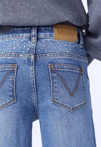 Jean en denim avec un délavage bleu clair, présentant une étiquette brune "Molo", des clous en argent décoratifs sur les poches arrière et des détails de couture standards.