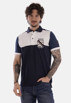 Uomo con capelli ricci e tatuaggi che indossa una polo blu navy e bianca con toppe, in piedi con una mano in tasca contro uno sfondo semplice.