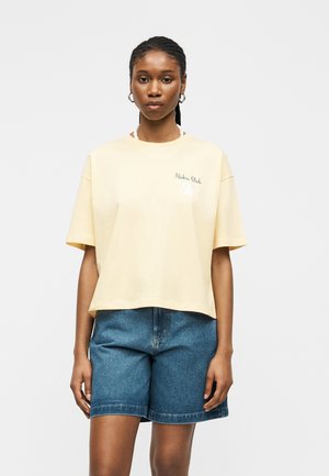 Femme portant un T-shirt jaune clair avec le texte "Aloha Club" et un short en jean bleu, debout devant un fond blanc uni.
