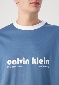 Blue cotton t-shirt with a white ribbed crew neck, featuring bold white text: "calvin klein," "West 39th Street," and "New York City."