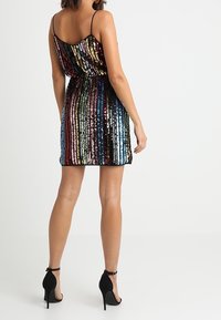 Frau in einem ärmellosen, mehrfarbigen, vertikal gestreiften Paillettenkleid mit Mini-Länge und schwarzen, hochhackigen Schuhen mit Knöchelriemen, mit dem Rücken zur Kamera.