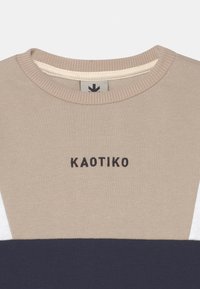 Beige cotton sweatshirt with a ribbed crew neck, featuring a graphic "KAOTIKO" logo in black and color-blocked sections in navy and white.