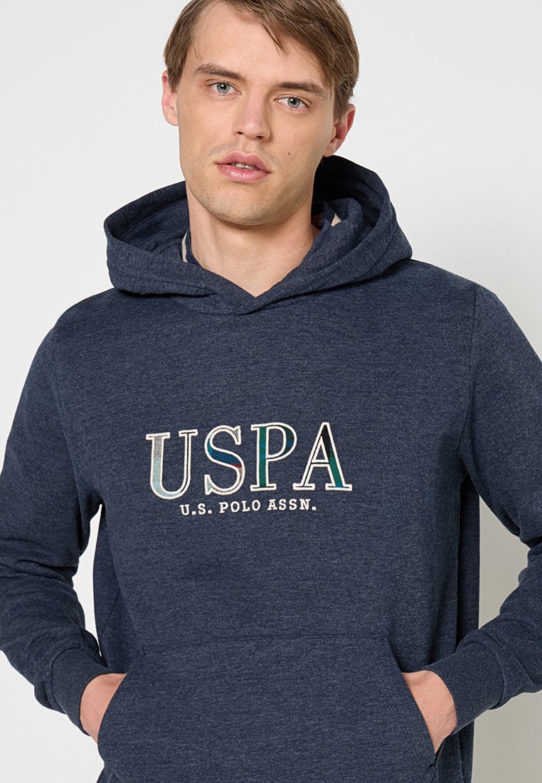Jeune homme portant un sweat à capuche bleu marine avec le logo "USPA U.S. Polo Assn.", les mains dans la poche avant, regardant légèrement sur le côté.