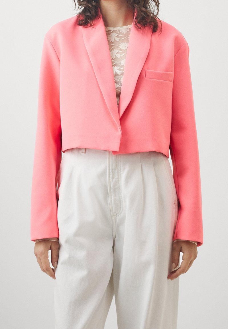 Blazer accorciato in rosa brillante, realizzato in tessuto morbido. Presenta una rever a punta, una tasca singola e maniche lunghe, abbinato a pantaloni bianchi a vita alta.